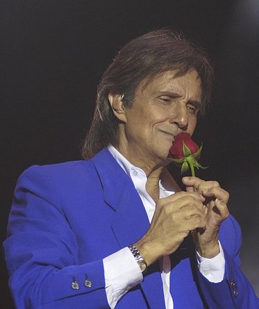 Roberto Carlos em concerto. Ele segura uma rosa próxima de seu rosto.