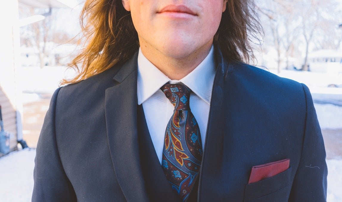 Homem com cabelo comprido vestindo paletó azul escuro, camisa branca e gravata azul estampada