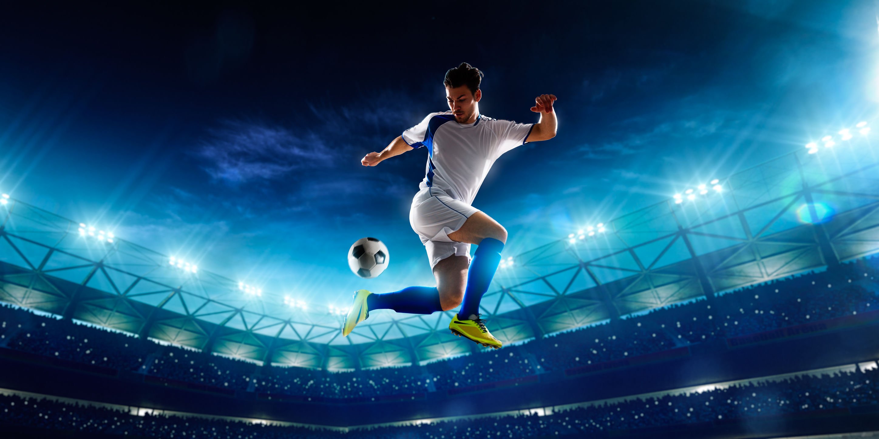 Jogador de futebol driblando bola no ar, e ao fundo a arquibancada do estádio acesa.