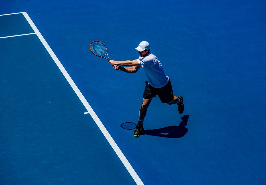 Homem jogando tênis em quadra azul, usando camiseta, shorts e um boné branco.
