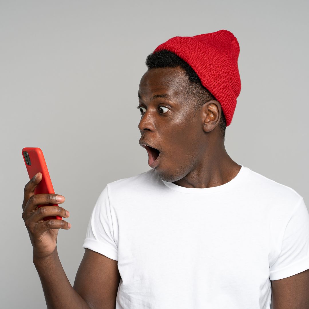 Homem negro segurando o celular enquanto olha para ele com uma expressão de assustado.