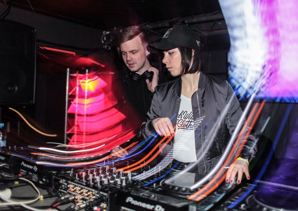 Homem e mulher trabalhando como DJ's em uma mesa de som de uma festa, cercados por feixes de luz em movimento.