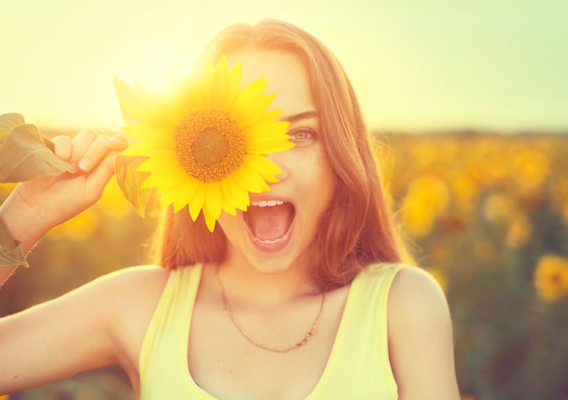 Garota com girassol em frente ao rosto e campo de girassol ao fundo com sol reluzindo