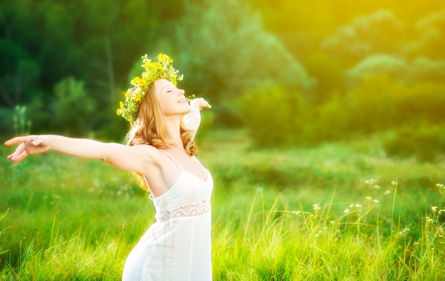 Mulher com braços abertos e coroa de flor na cabeça com fundo ver ao fundo e luz do sol