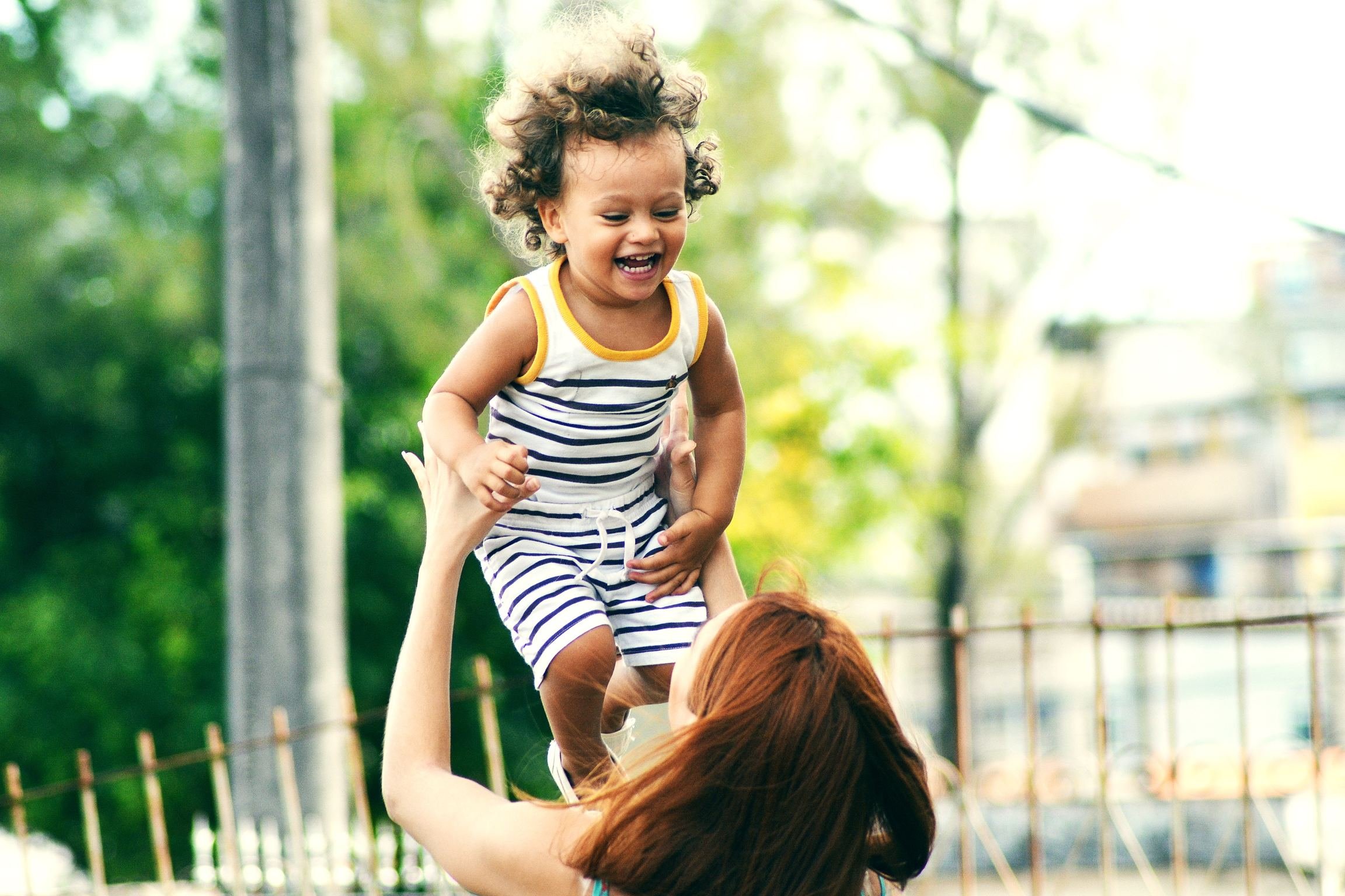 Mulher segurando criança pequena para o alto, que sorri e olha para ela.