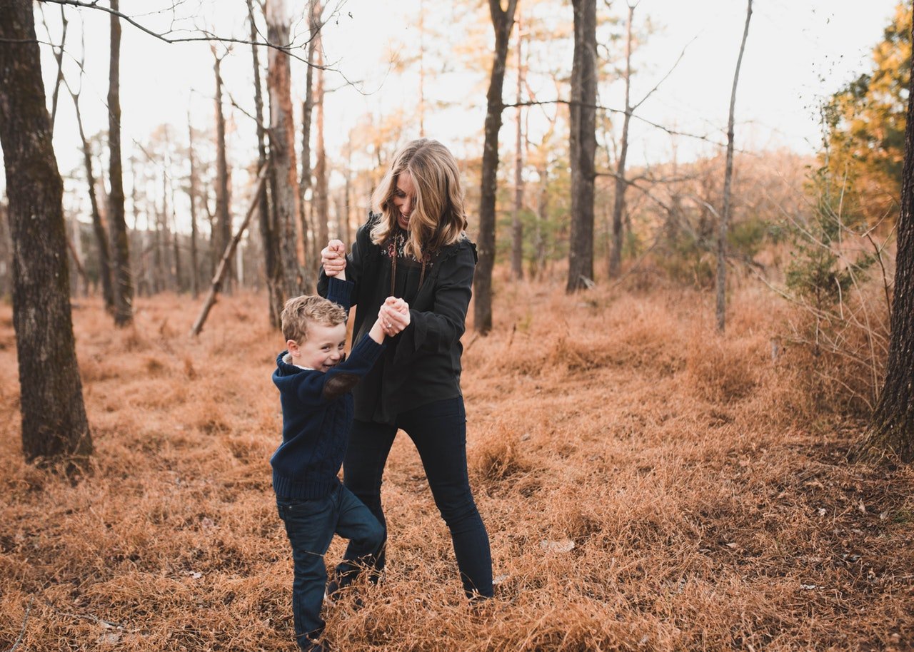 Menino pequeno se apoiando em mulher, que o segura pelos braços enquanto ele tenta andar. Eles estão em um campo com grama marrom e árvores sem folhas.