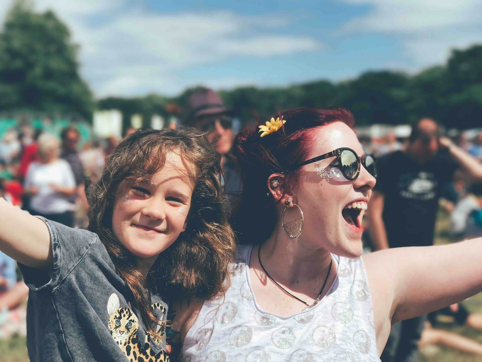 Menina e mulher lado a lado, de braços abertos e sorrindo, durante um festival.