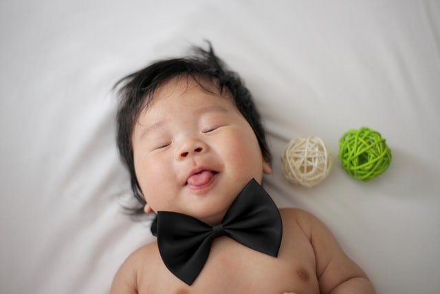Bebê deitado com gravata borboleta e duas bolinhas de lã ao lado de olhos fechadinhos