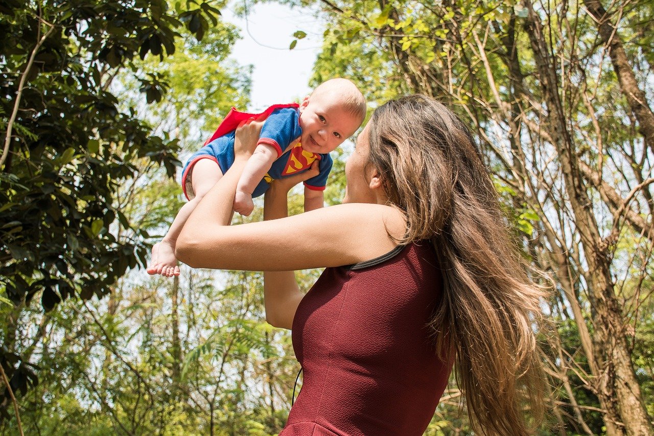 Mulher de costas segurando um bebê vestido de super homem no alto