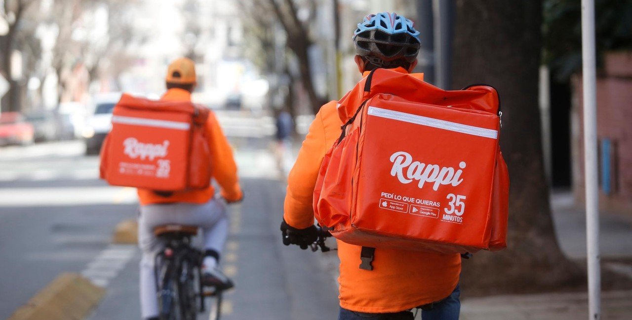 Dois entregadores da Rappi, um usando capacete e o outro boné, andando de bicicleta com a mochila de entrega da empresa.