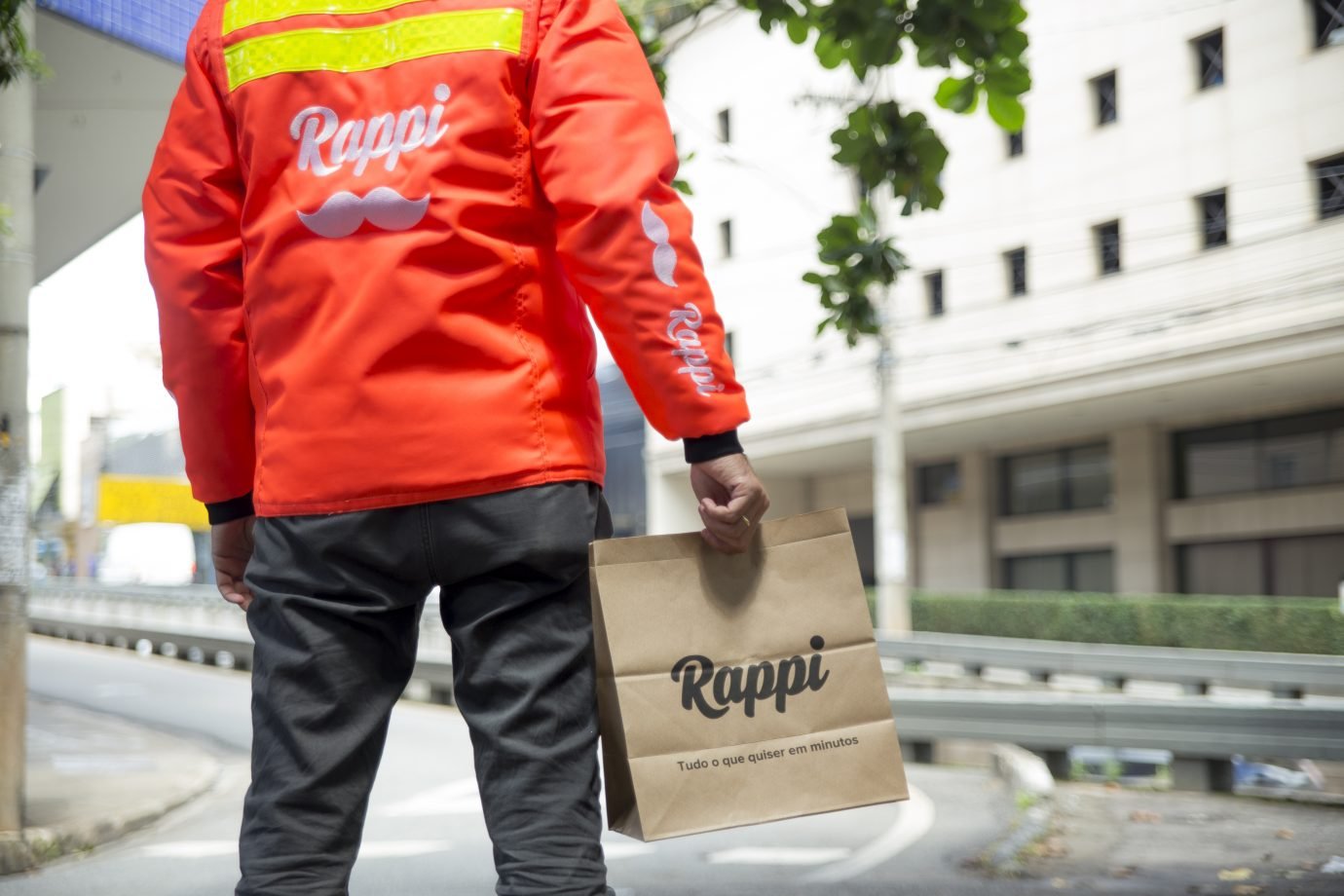 Entregador da Rappi com jaqueta da empresa segura uma sacola de papel, com o logo da Rappi.