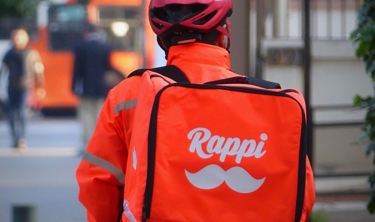 Entregador da Rappi com jaqueta e mochila de entrega da empresa, e capacete vermelho, de costas para a câmera.