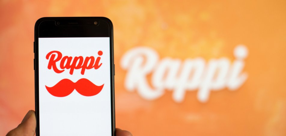 Celular com tela em branco e o logo da Rappi, e ao fundo uma parede laranja com 'Rappi' escrito em branco.