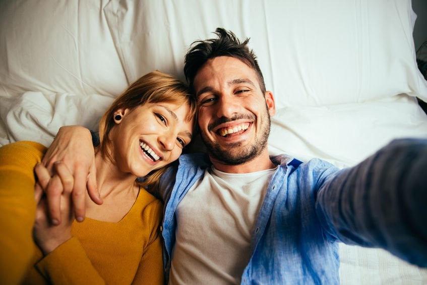 Jovem casal deitados na cama abraçados tirando selfie.