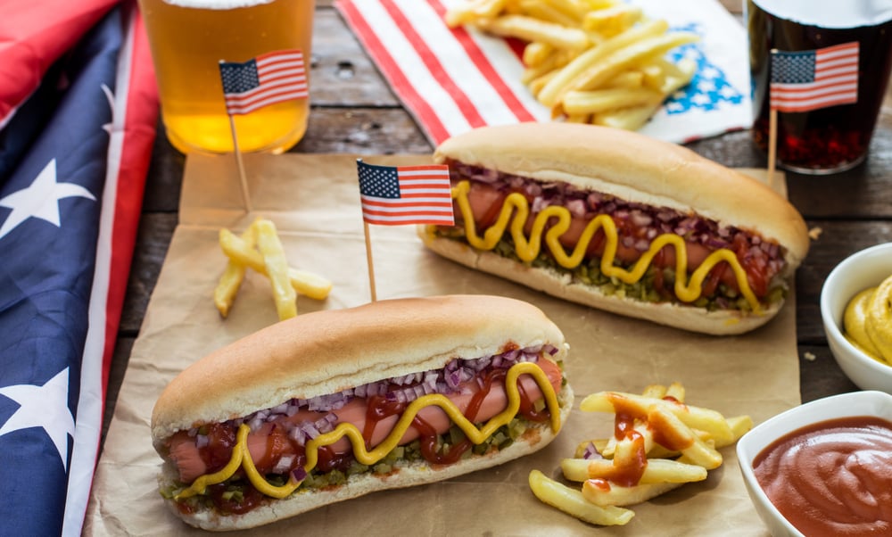 Cachorros-quentes com mini-bandeiras dos Estados Unidos. Há batata frita e refrigerante no tabuleiro