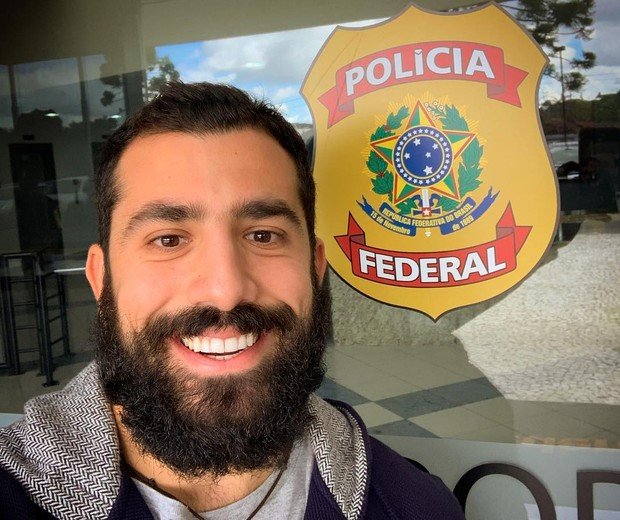Homem sorrindo ao lado de um símbolo da polícia federal
