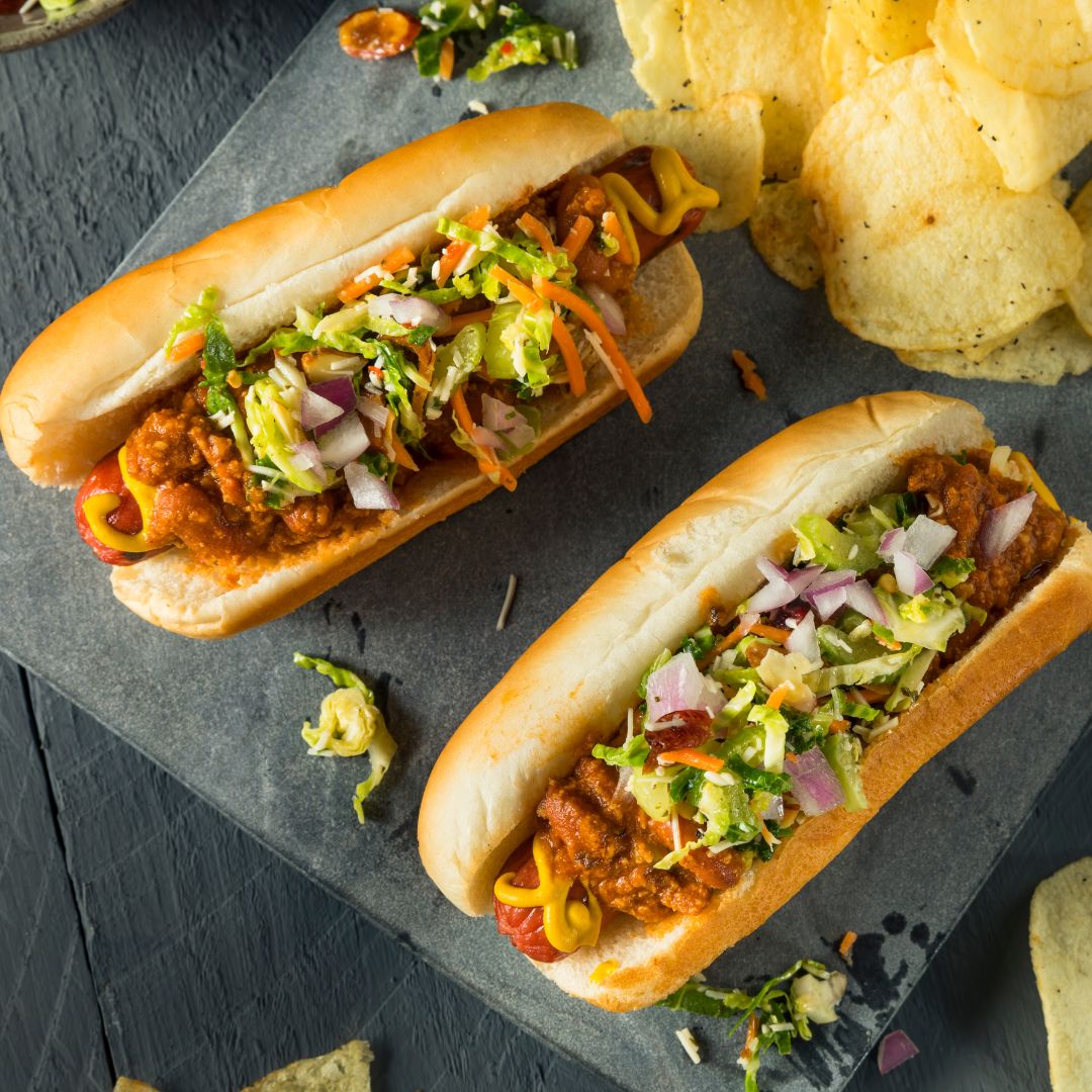 Imagem de dois hot dogs com bastante ingredientes e alguns chips ao lado