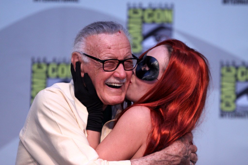 Stan Lee sorridente recebendo beijo na bochecha de mulher cosplayer