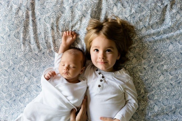 Menina branca deitada ao lado de um bebê branco, ambos com roupas brancas