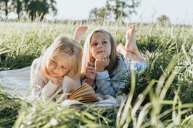 Duas garotas brancas e loiras deitadas na grama lendo um livro