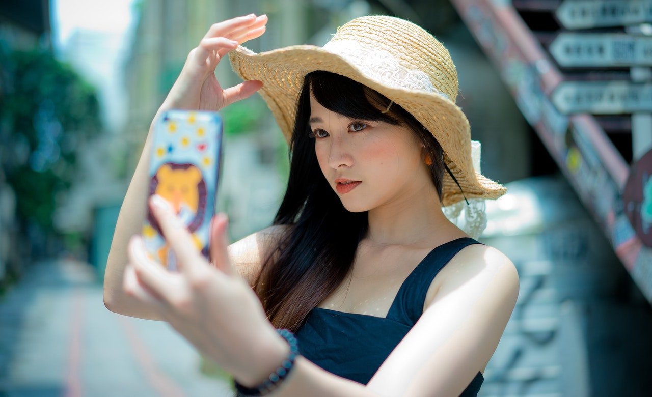 Mulher tira selfie em ambiente externo. Ela usa um chapéu e segura a aba dele com os dedos.