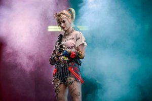 Divulgação Warner Bros Personagem Arlequina com fundo de fumaça azul e rosa apontando arma para frente