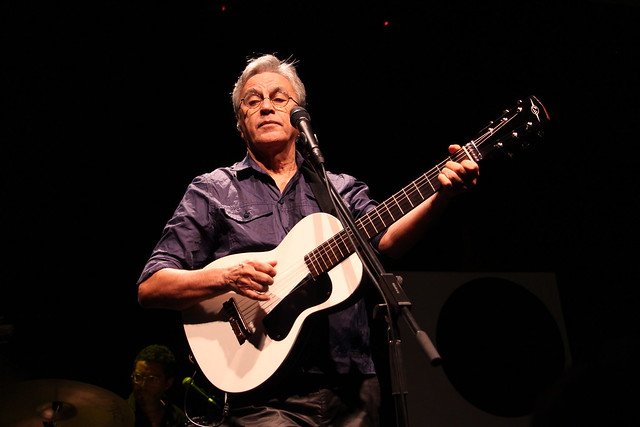 Homem branco de cabelos grisalhos segurando violão branco.