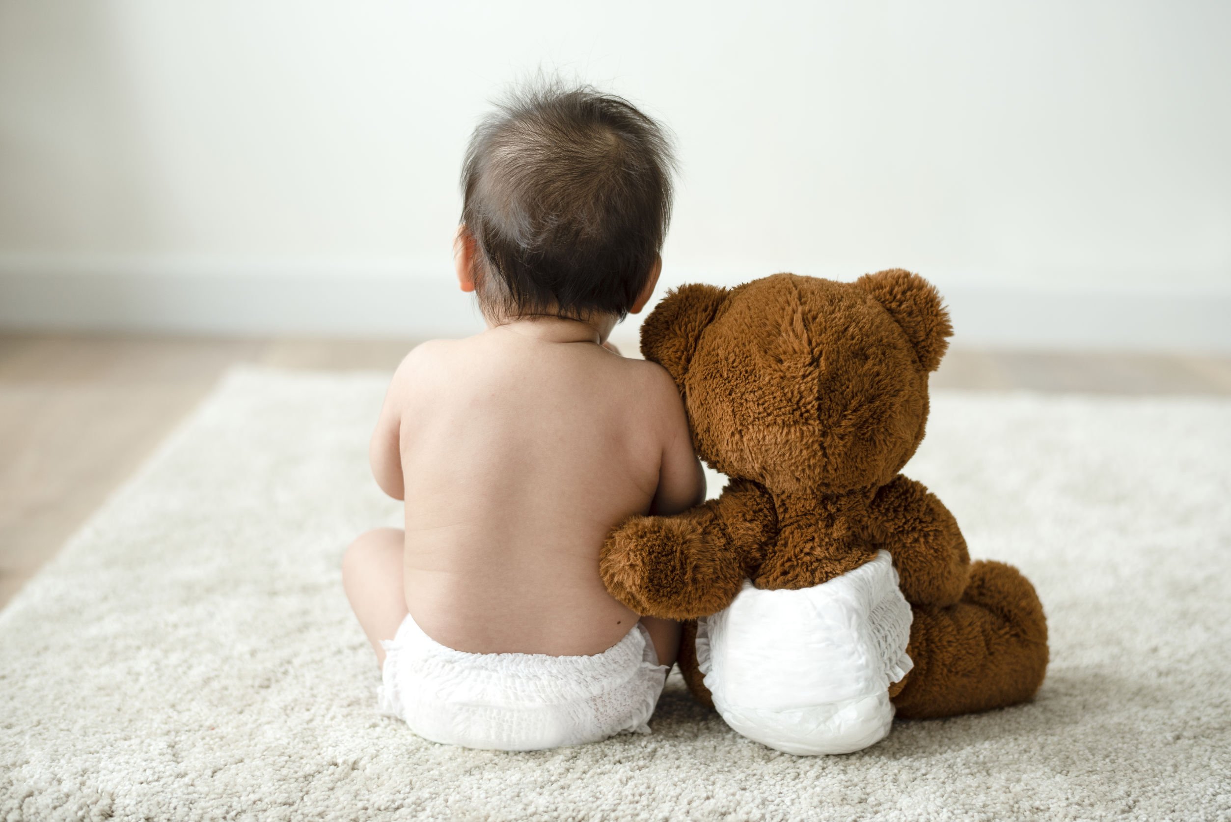 Bebê sentado no tapete ao lado de um ursinho de pelúcia