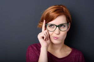 Mulher de óculos com dedo no rosto e expressão desconfiada