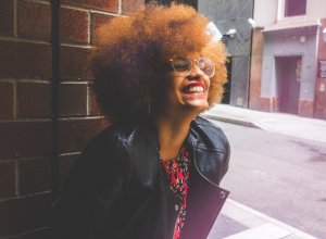 Mulher com black power e óculos sorrindo apoiada numa parede