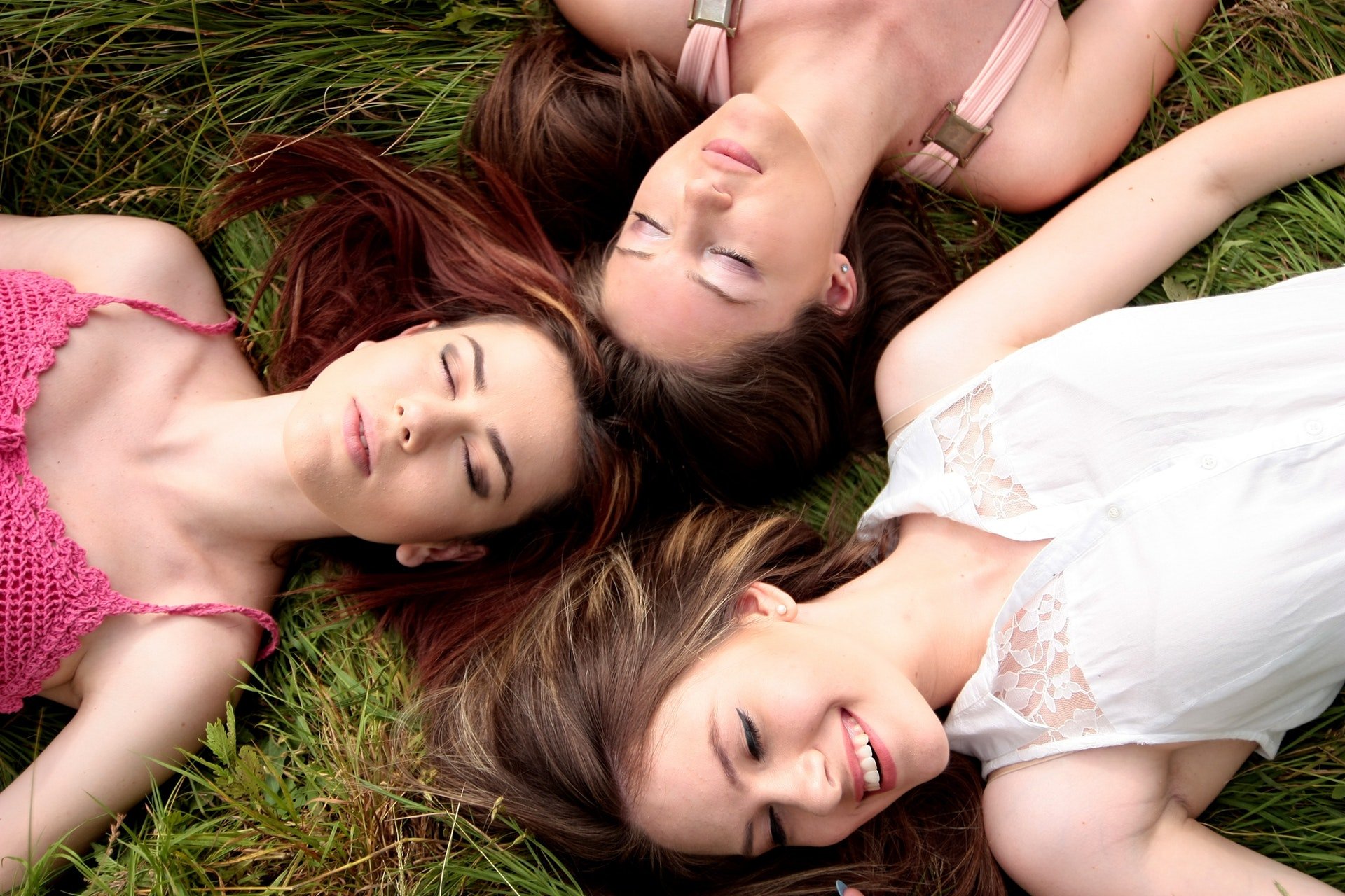 Três amigas deitadas com as cabeças próximas, de olhos fechados num gramado