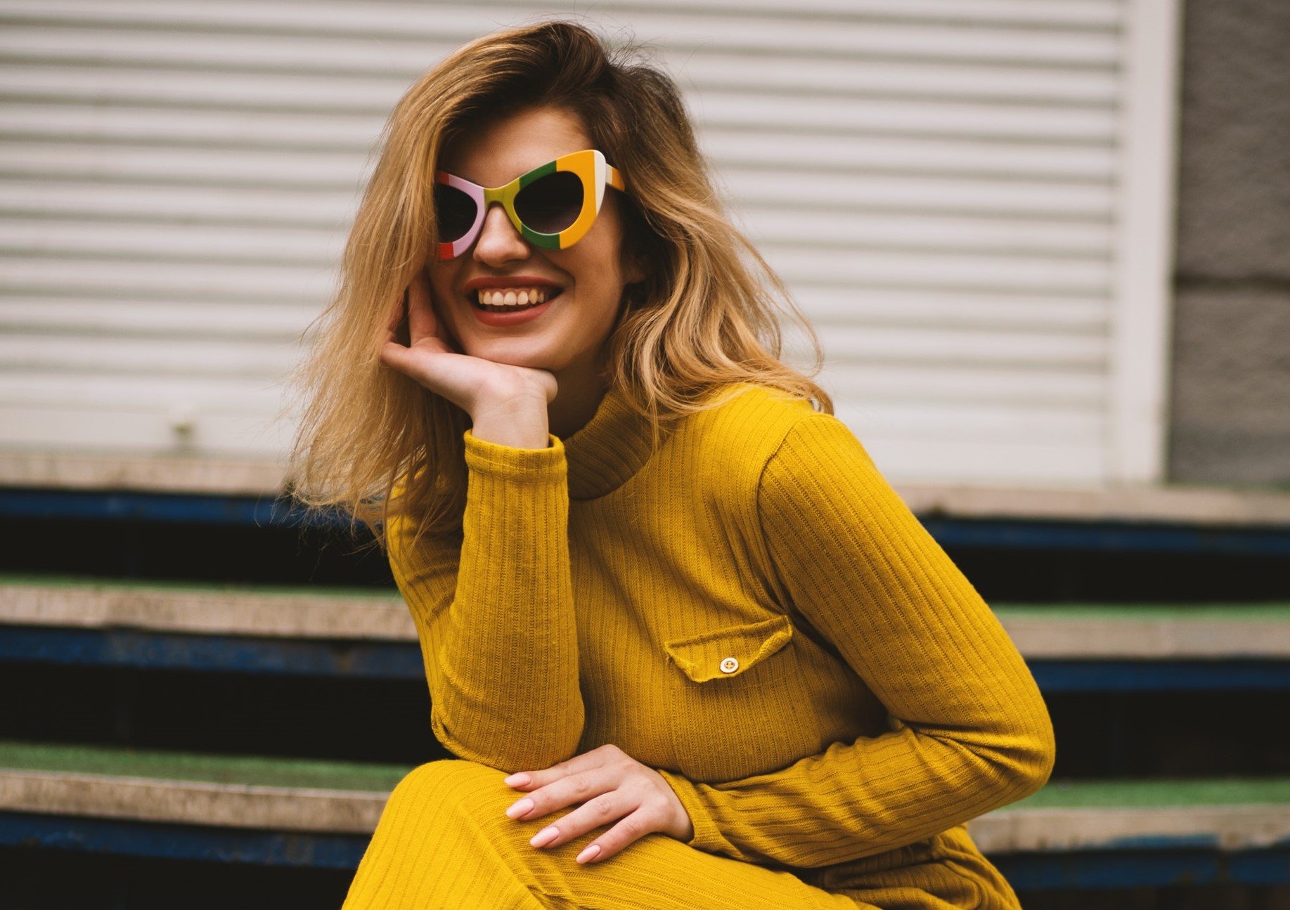 Mulher de óculos de sol e vestido amarelo sorrindo com a mão apoiada no rosto