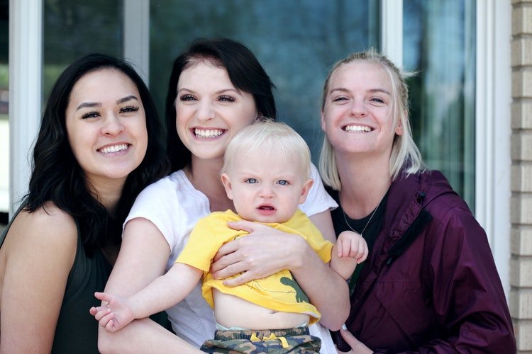 Família com três mulheres sorrindo, a mulher do meio segura um bebê