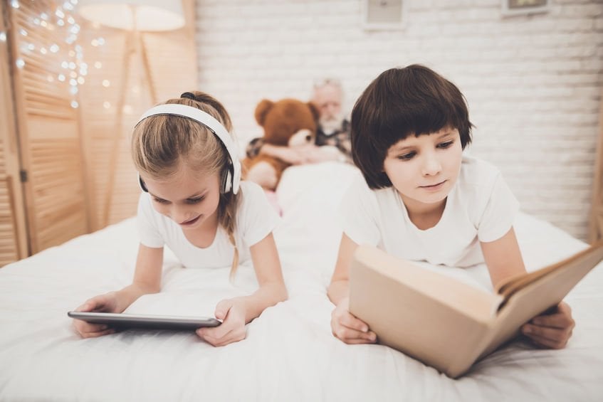 Criança deitada olhando um tablet, enquanto outra lê um livro