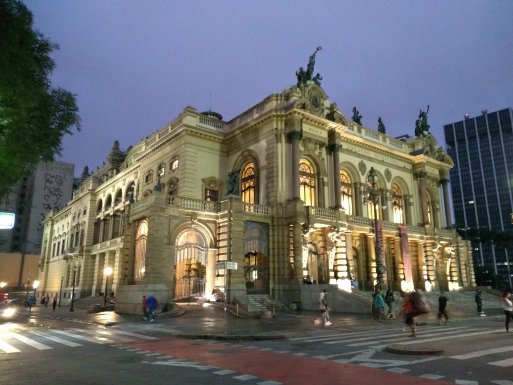 Fachada do Teatro Municipal de São Paulo, com céu ainda claro e luzes acesas.