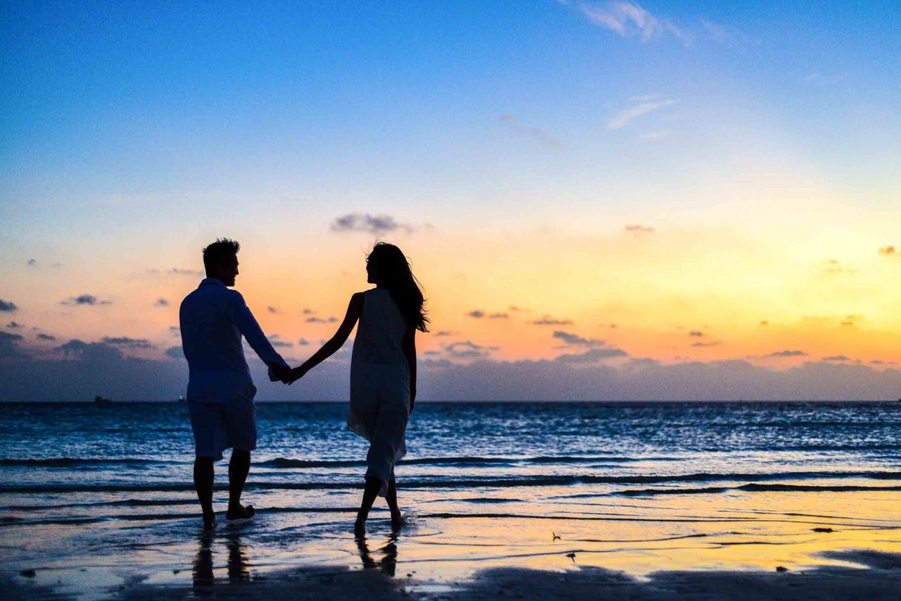 Homem e Mulher andando de mãos dadas na praia durante o pôr do sol.