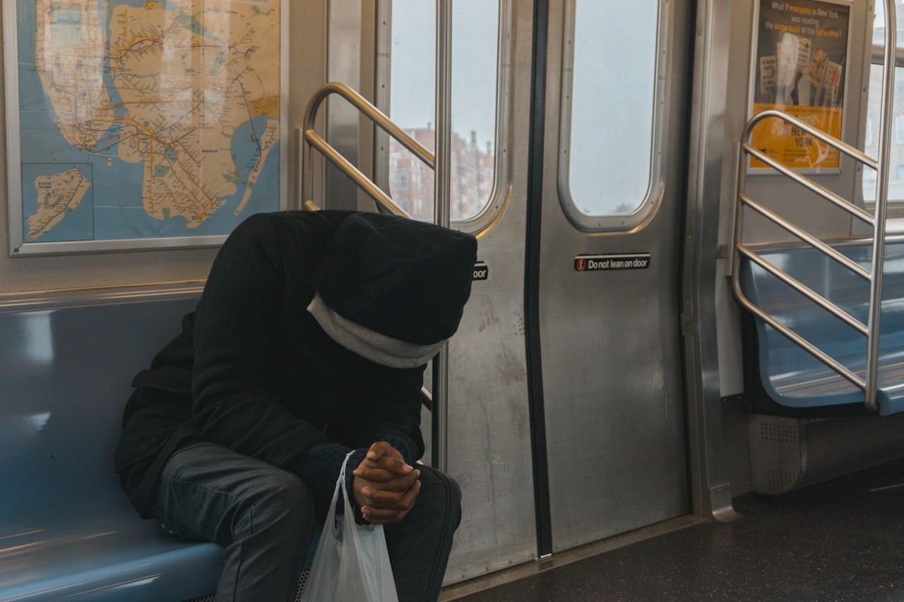 Pessoa sentada no metrô, usando capuz e de mãos dadas com a cabeça baixa.