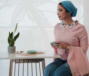 Mulher sentada em frente de uma mesa, segurando uma xícara. Vestindo blusa comprida e um lenço na cabeça.