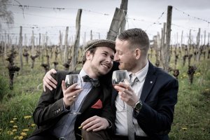 Dois homens se abraçando e segurando taças de vinho. Ambos usam ternos, e estão em algum lugar no campo.