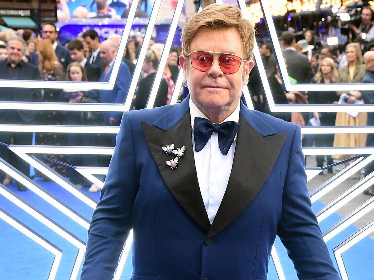 Elton John em evento, com terno azul e gravata borboleta grande da mesma cor