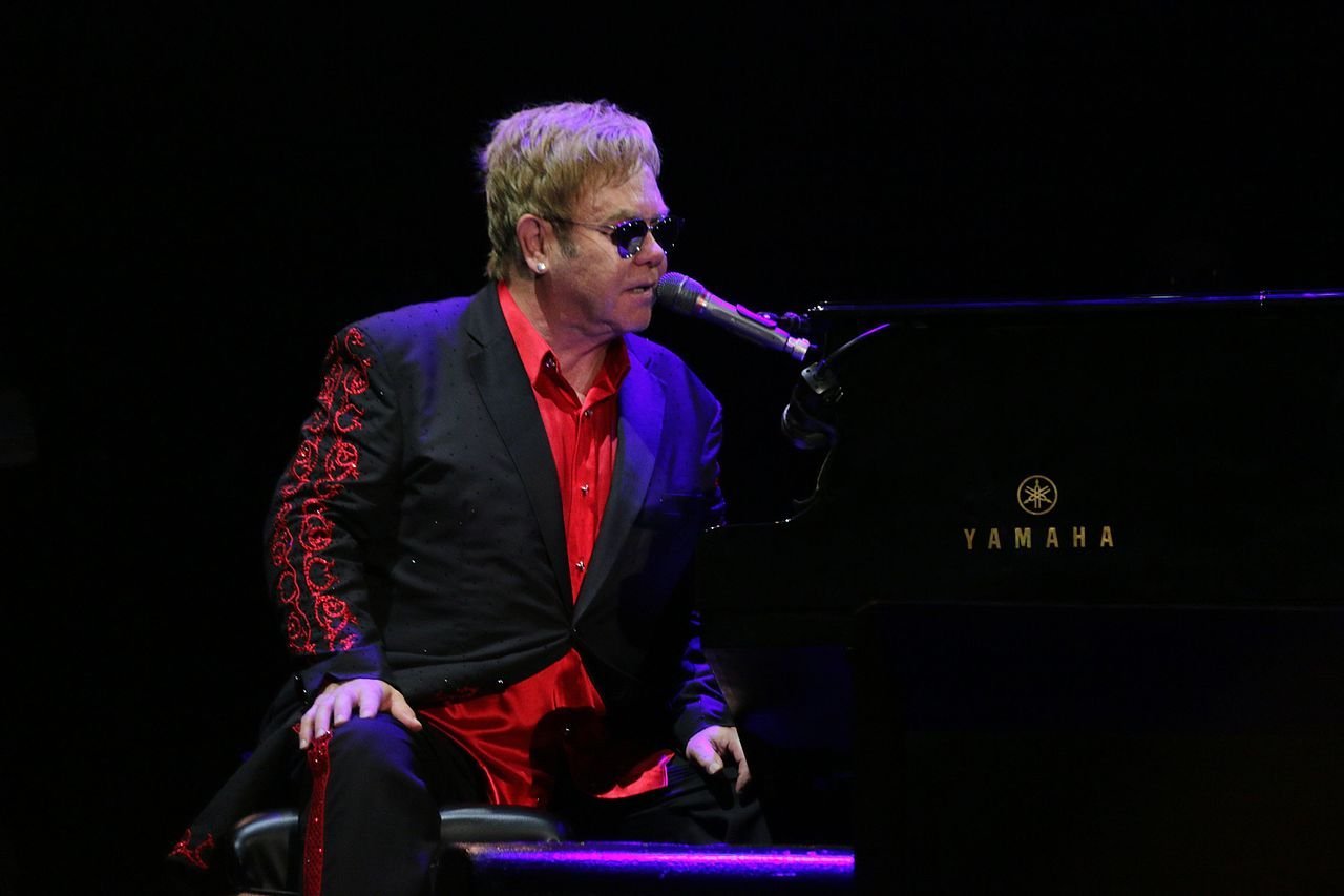 Elton John em show usando terno preto com camisa vermelha por baixo