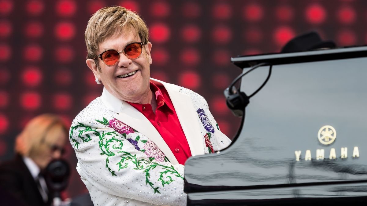 Elton John tocando piano em show, usando terno branco com bordados e camisa vermelha