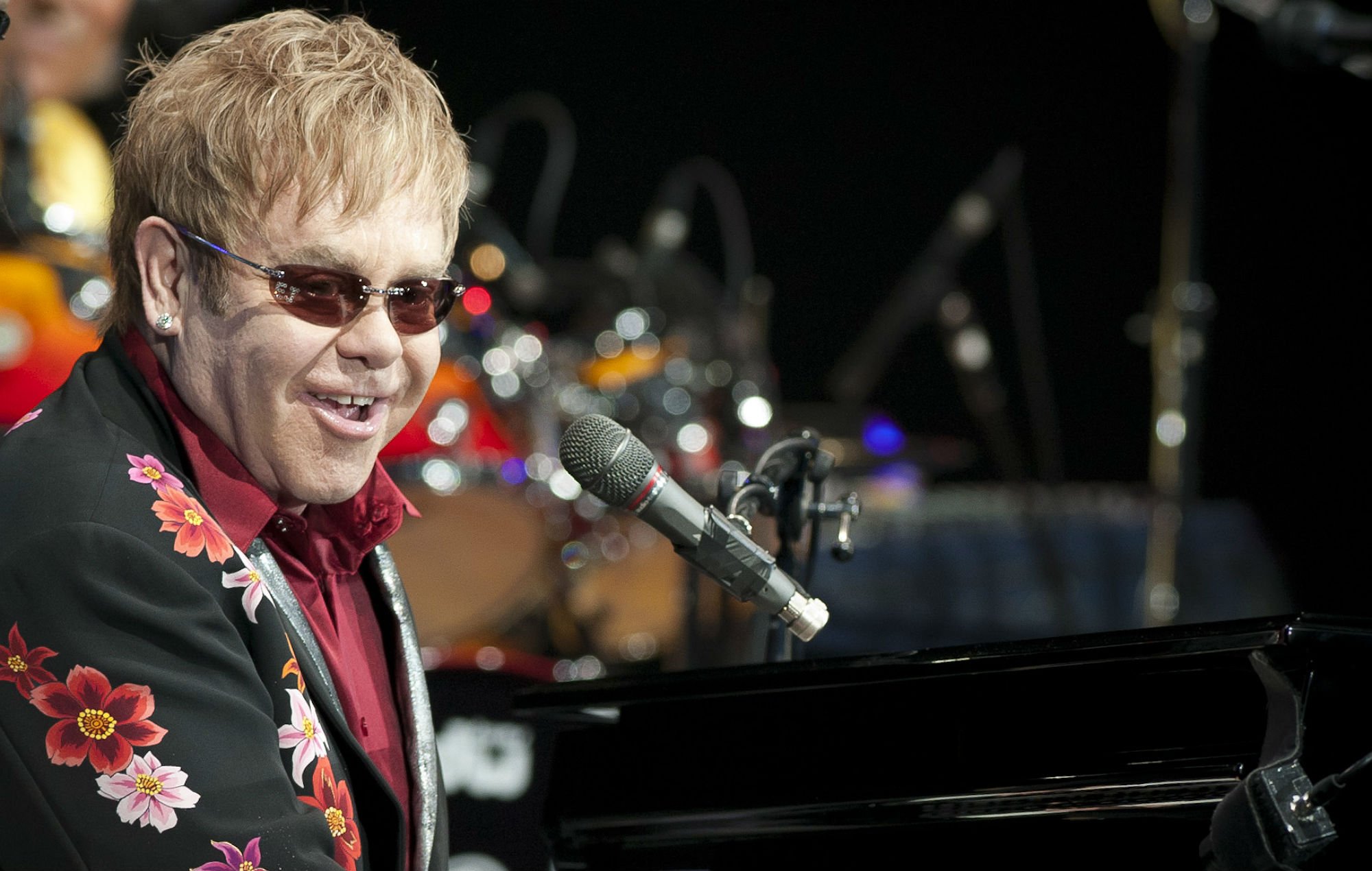 Elton John em show, usando terno preto com desenhos de flores e camisa vermelha