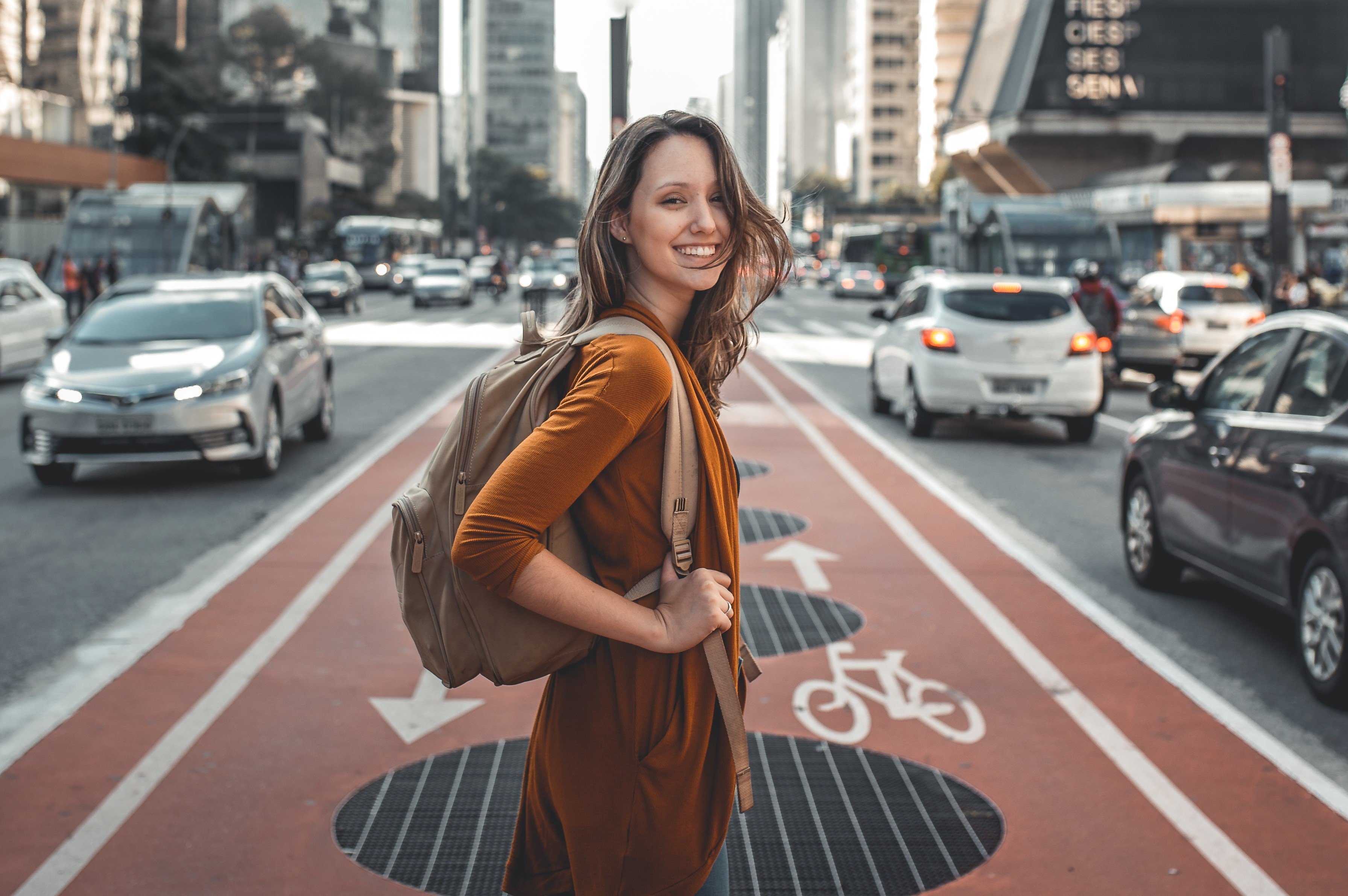 Mulher de mochila, sorrindo, no meio de uma avenida movimentada