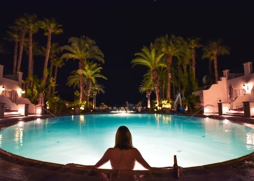 Mulher de costas sentada na beirada de uma piscina, à noite.