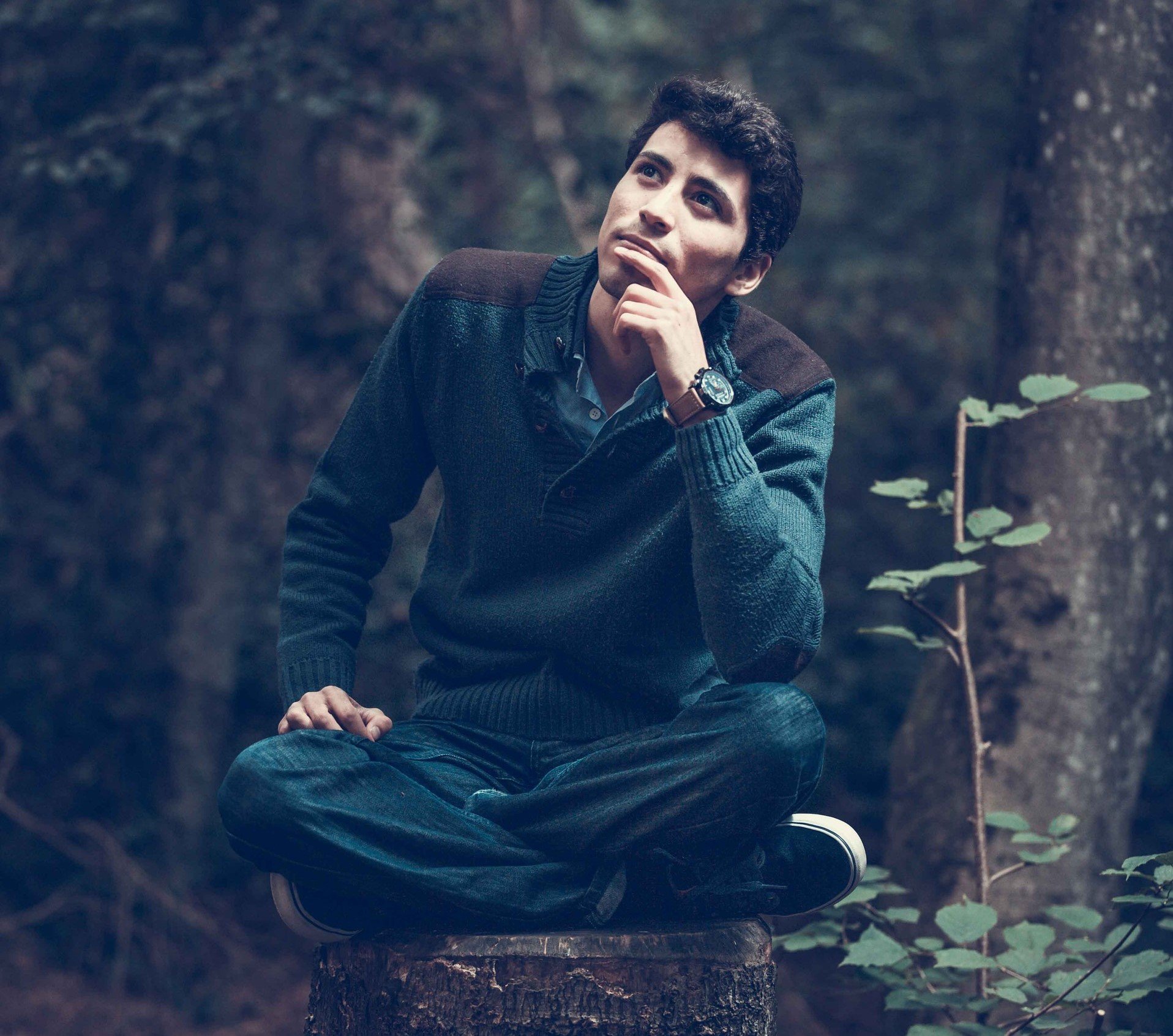 Homem sentado numa floresta com a mão no rosto, pensando