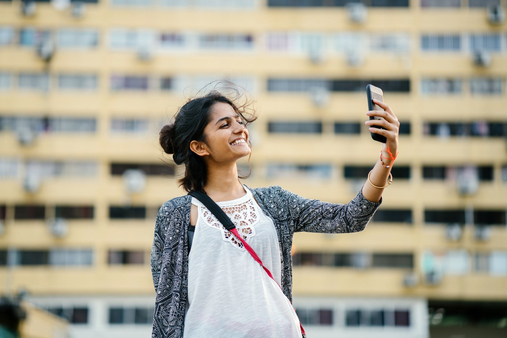 Mulher em frente a prédio, tirando selfie e sorrindo