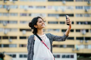 Mulher em frente a prédio, tirando selfie e sorrindo