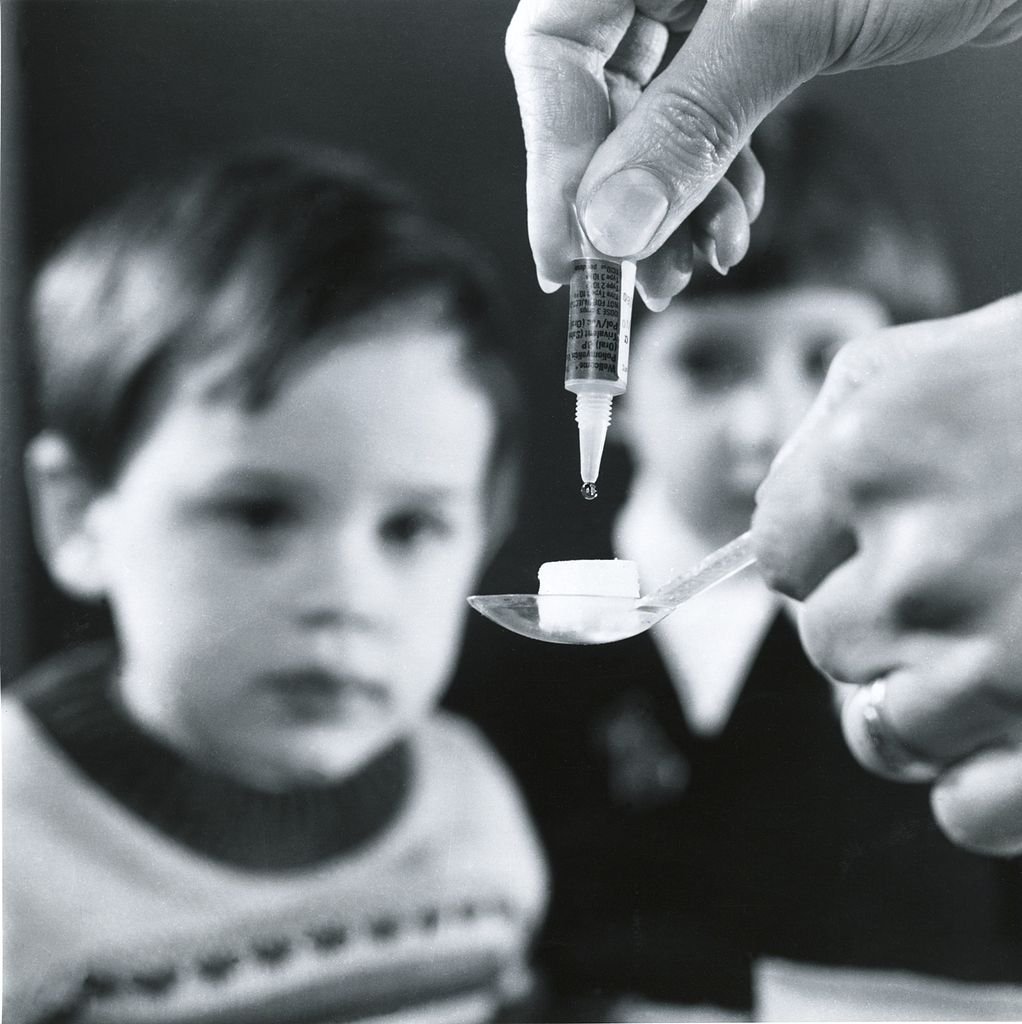 Menino esperando para tomar vacina contra poliomielite. Uma pessoa pinga a vacina em uma colher junto a um cubo de açúcar.
