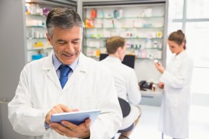 Farmacêutico usando tablet, enquanto atrás dele outra farmacêutica conversa com cliente.